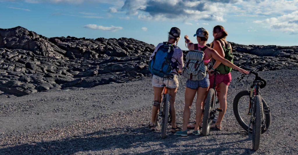 Three women with backpacks stand next to their bikes on Chain of Craters Road at Hawaii Volcanoes National Park.